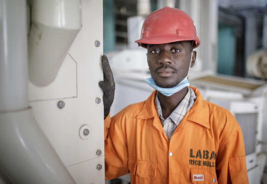 Bild mit einem Mann mit Helm und orangen Arbeitsoverall neben einem großen Maschinengehäuse. Als Beispiel für eine internationale Fachkraft für Industriemechanik.